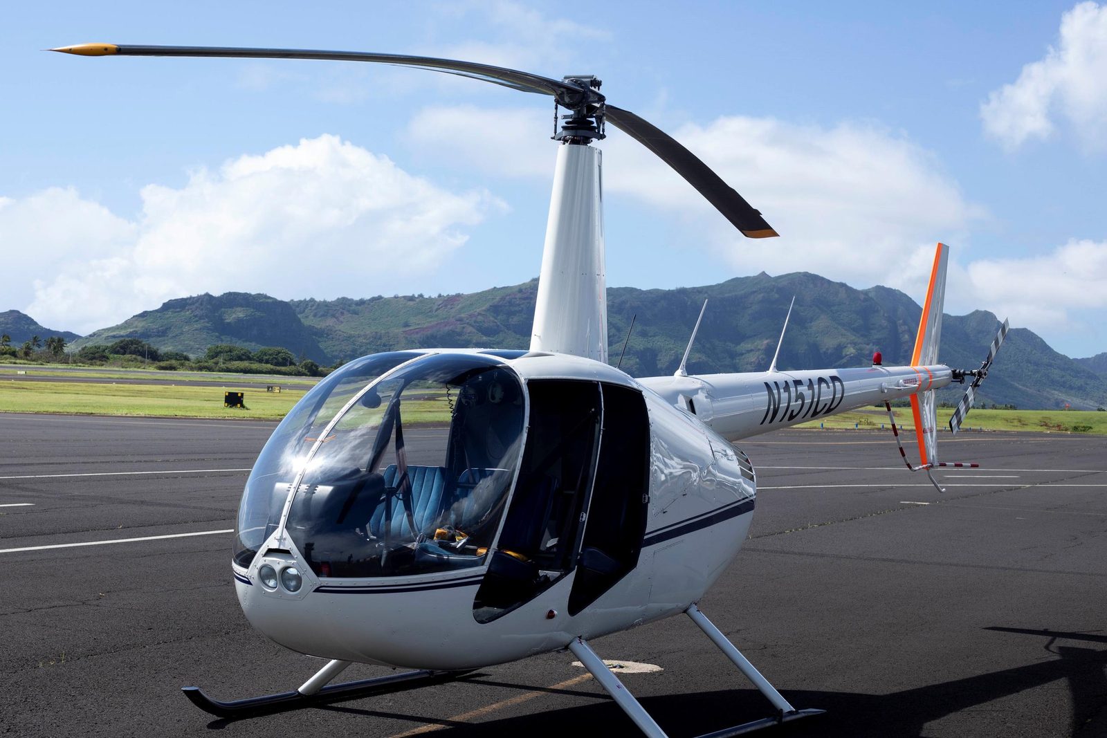 A white four-seat helicopter parked on the ramp at Līhuʻe Airport with green hills behind