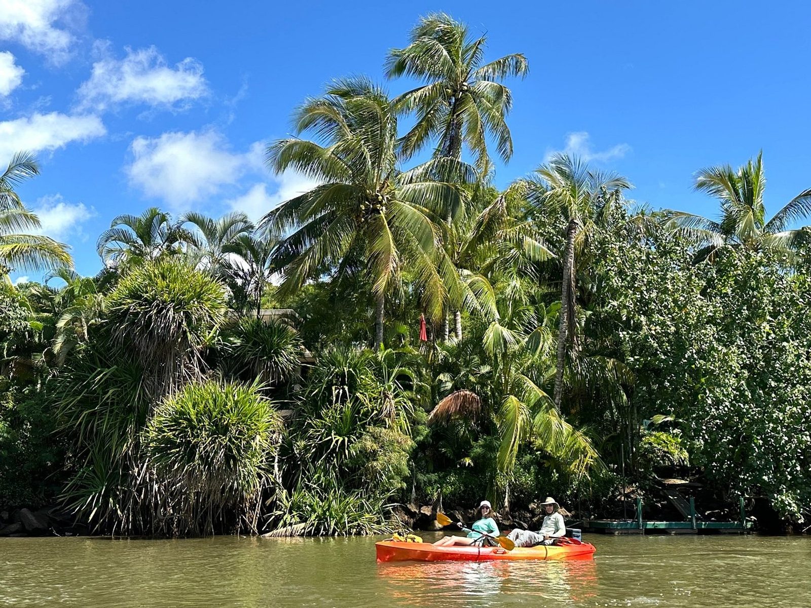 Two guests in a red tandem kayak paddling the calm Wailua River under a wall of palms and jungle