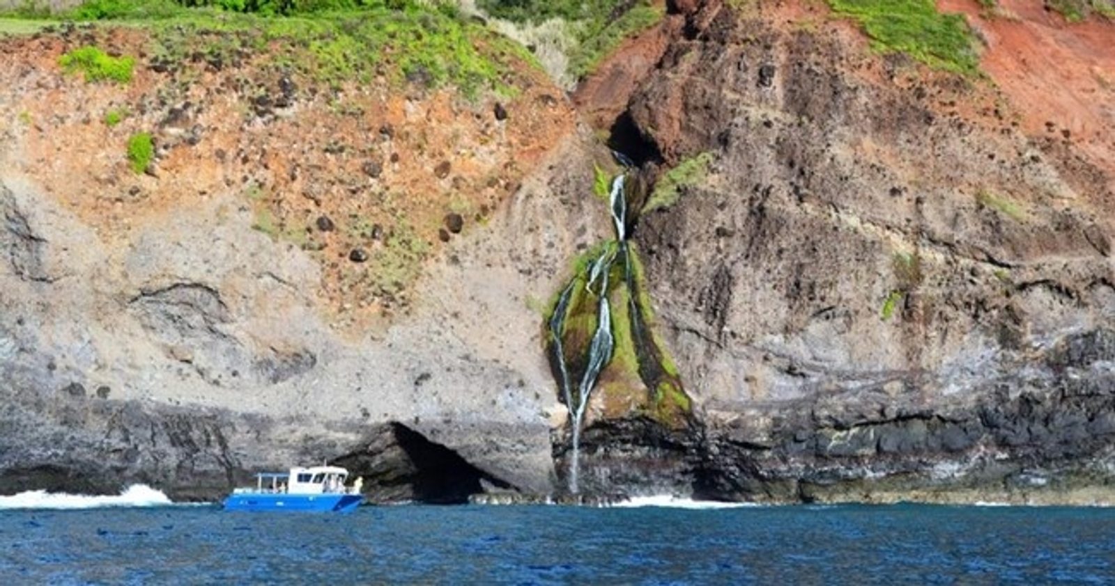 The catamaran idling below a waterfall that threads down a red and green cliff into a sea cave