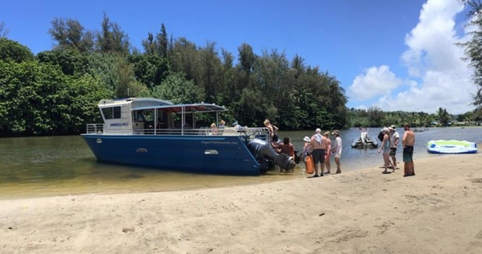 Guests wading out to the catamaran from the sand at Hanalei Bay under ironwood trees
