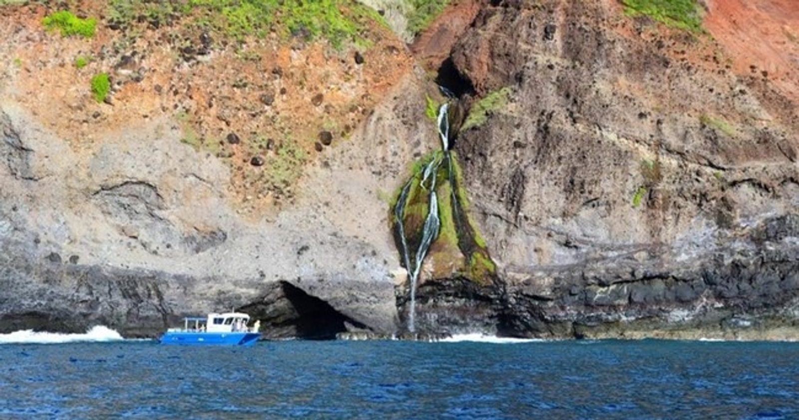 A thin waterfall dropping into a sea cave at the waterline with the boat small beside it