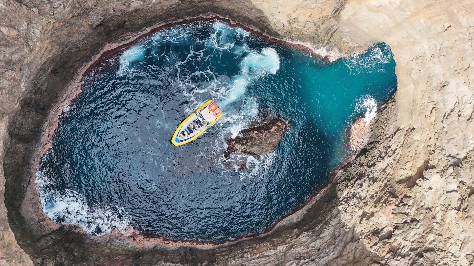Aerial view of the raft inside a round sea cove with a collapsed roof, blue water surrounded by rock walls