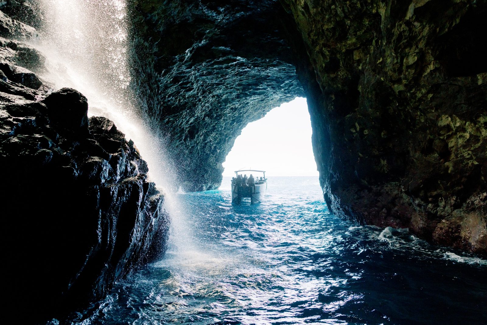 Looking out from inside a dark sea cave at a waterfall pouring over the cave mouth, with a tour boat waiting in the sunlit water beyond