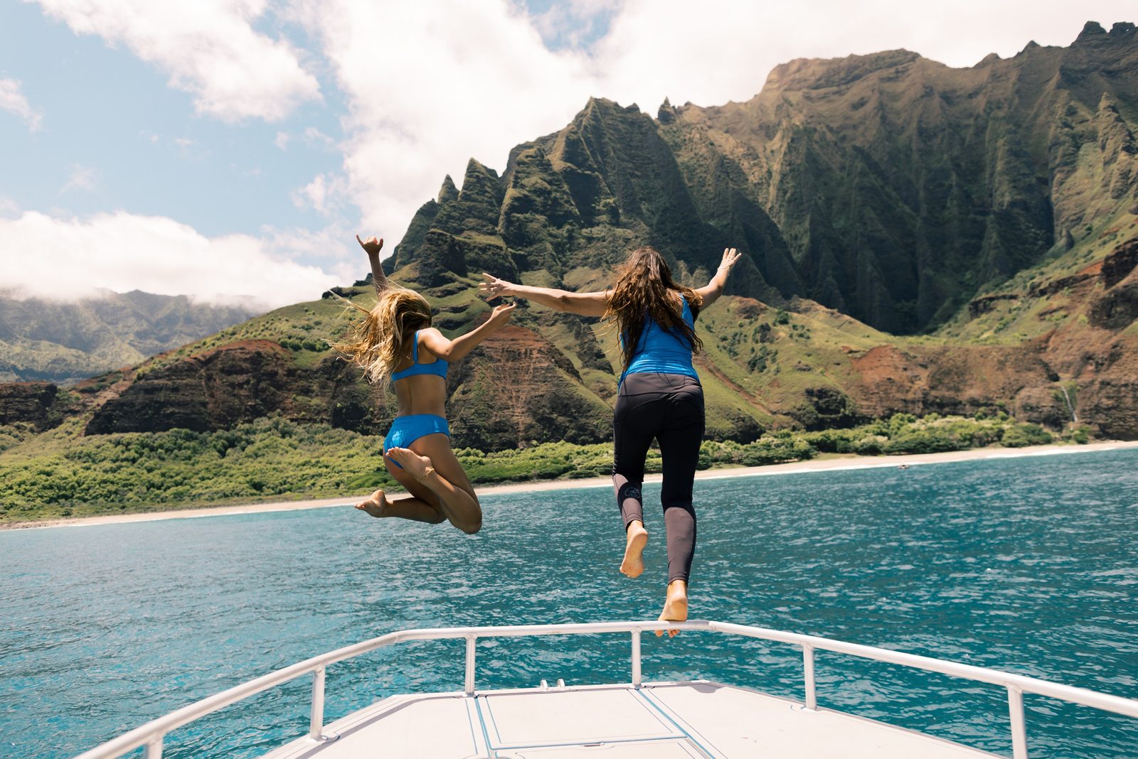 Two guests leaping off the bow of the catamaran into blue water with the Nā Pali cliffs rising behind them