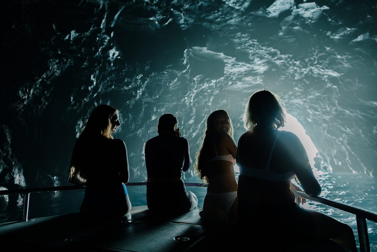 Four guests sitting on the bow rail inside a sea cave, silhouetted against the blue light at the cave entrance