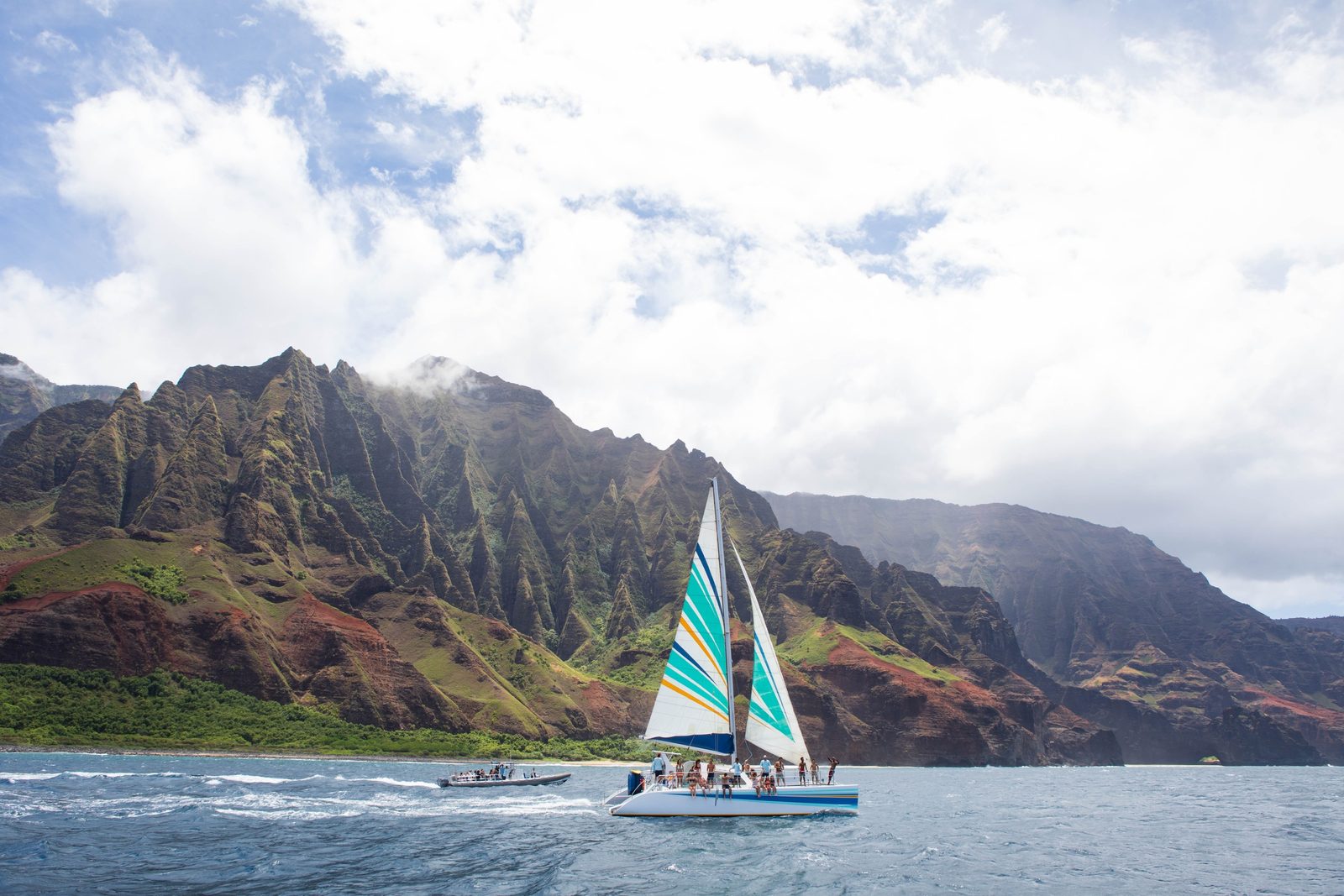 A sailing catamaran with teal and white sails running along the base of the green fluted Nā Pali cliffs