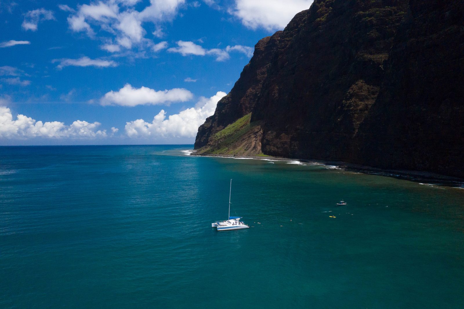 A small white sailboat dwarfed by a tall dark Nā Pali headland above deep blue water