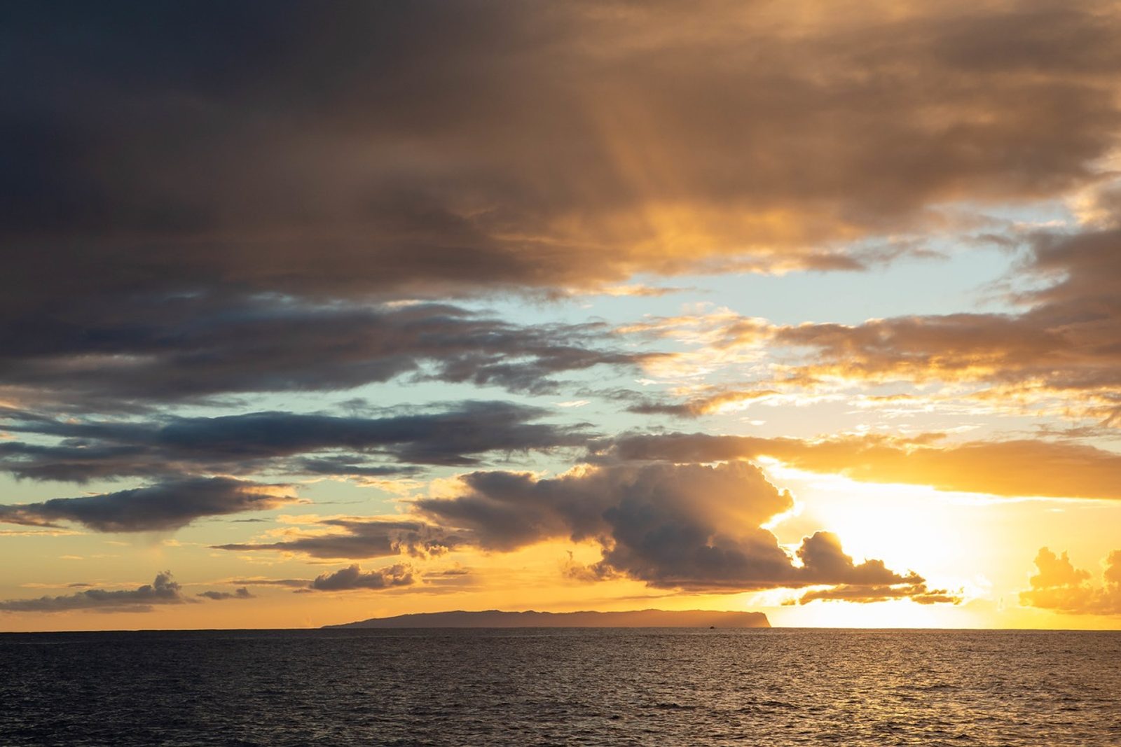 Golden rays breaking through dark clouds over the ocean and Niʻihau at sunset