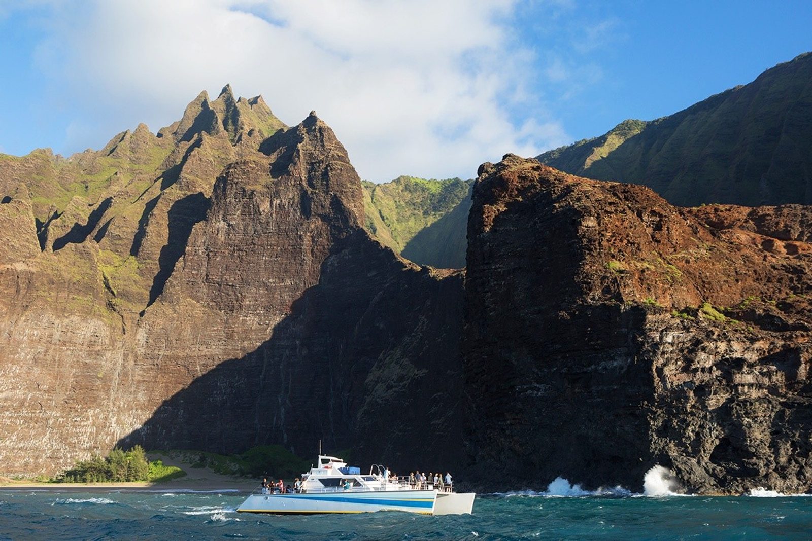 A white catamaran full of guests below sharp rock spires on the Nā Pali Coast on a sunny afternoon