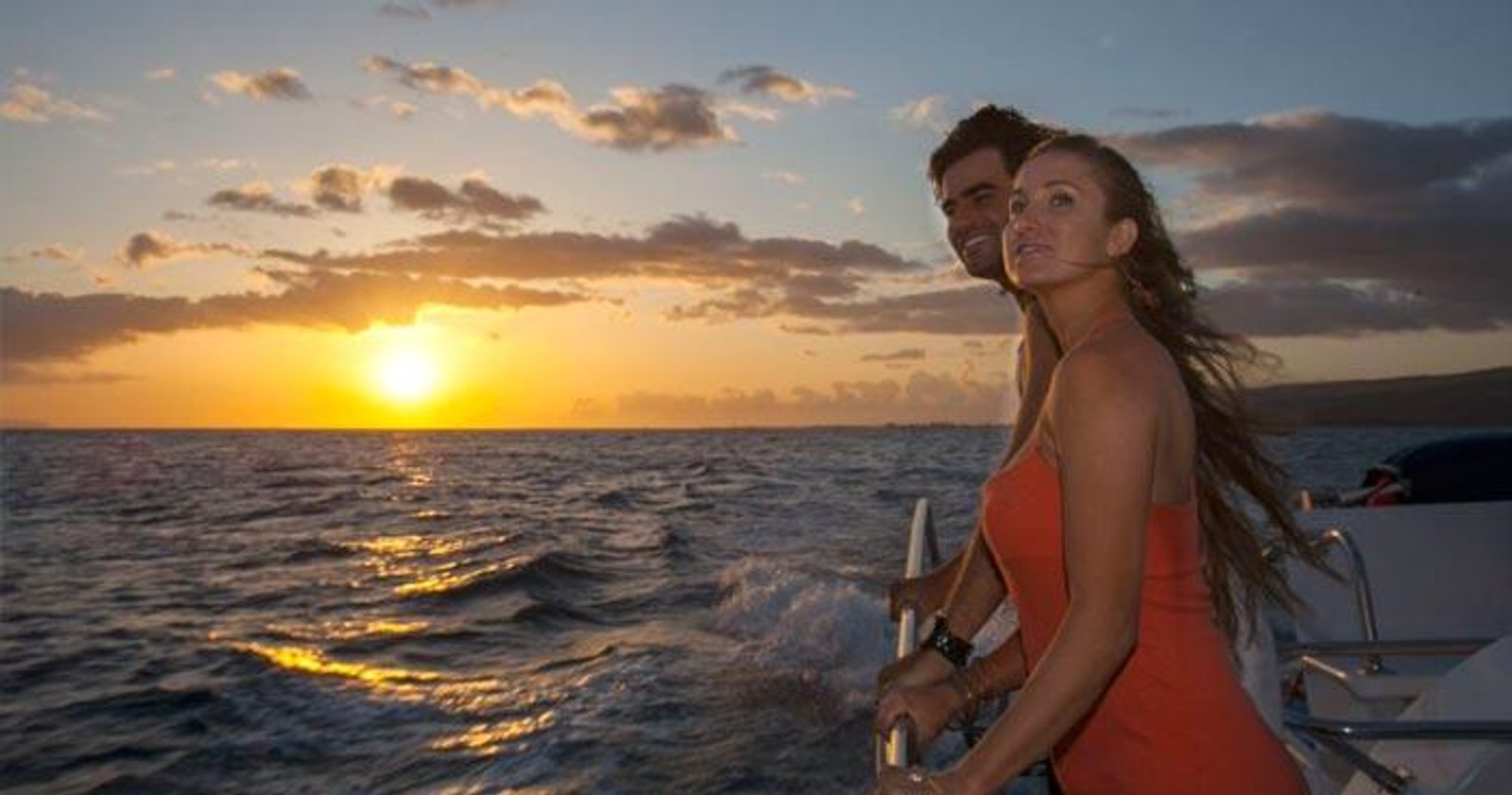 A couple at the rail of the boat with the sun setting over the water behind them