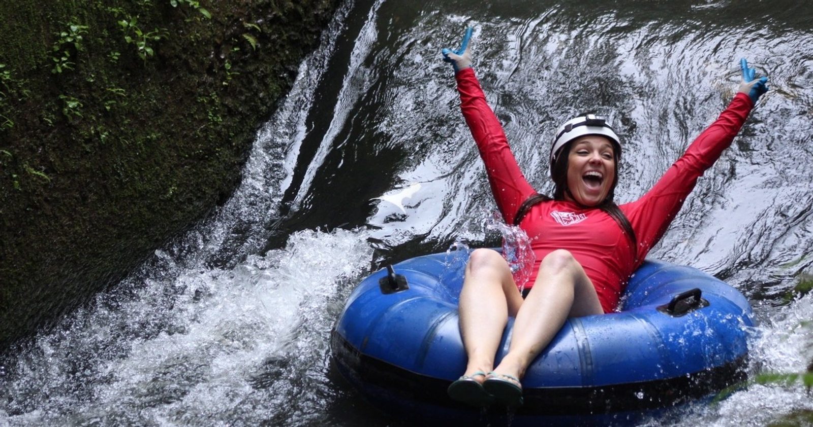 A woman in a red shirt and helmet with both arms out riding a tube through a narrow flume